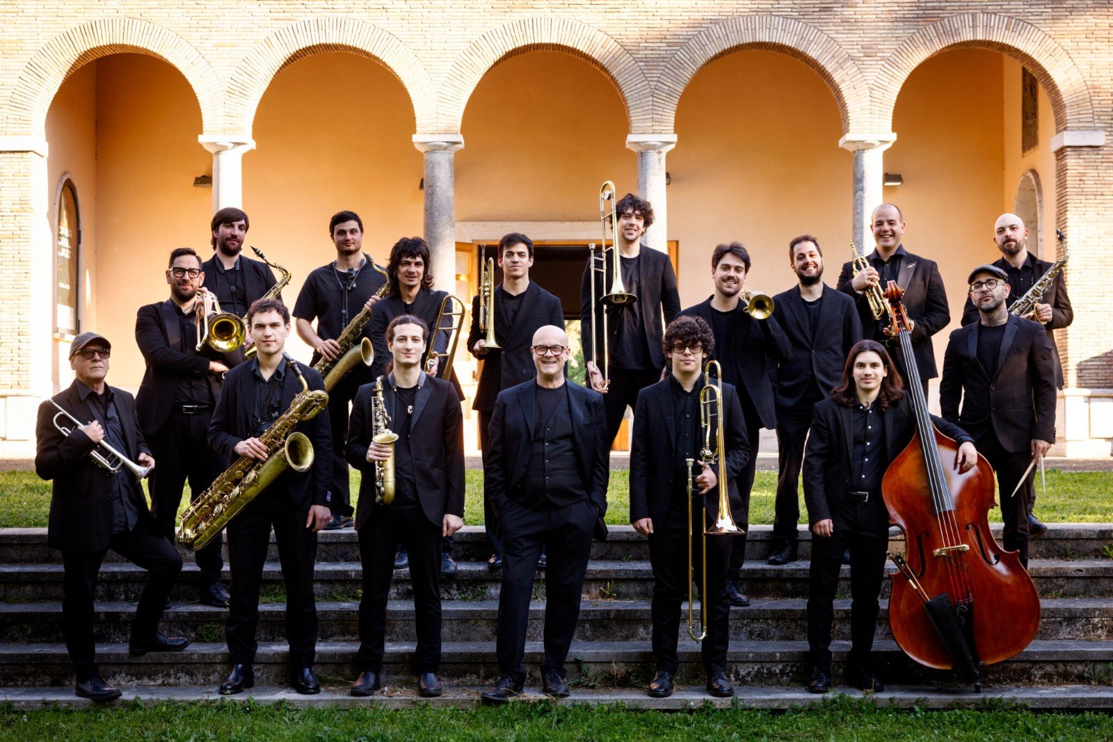 Foto di gruppo della Casa del Jazz Orchestra: musicisti vestiti di nero con sassofoni, trombe, tromboni e un contrabbasso, in piedi sui gradini davanti a un portico ad archi